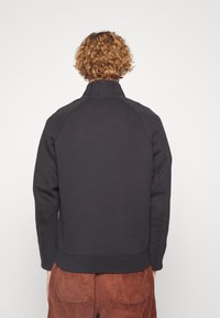 Sudadera negra con cuello alto, mangas raglán y dobladillo acanalado. La tela parece texturizada, combinada con pantalones de pana marrón.