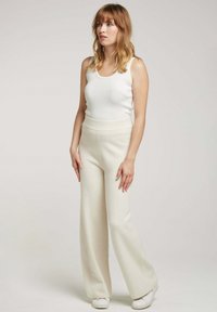 Pantalons larges de couleur crème en tissu doux, associés à un débardeur blanc ajusté, portés avec des baskets blanches ; design minimaliste, texture lisse.