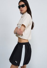 Fekete sportnadrág fehér csíkokkal, bézs, rövid ujjú pólóval párosítva. A modellen sötét napszemüveg van. Egyszerű háttér.