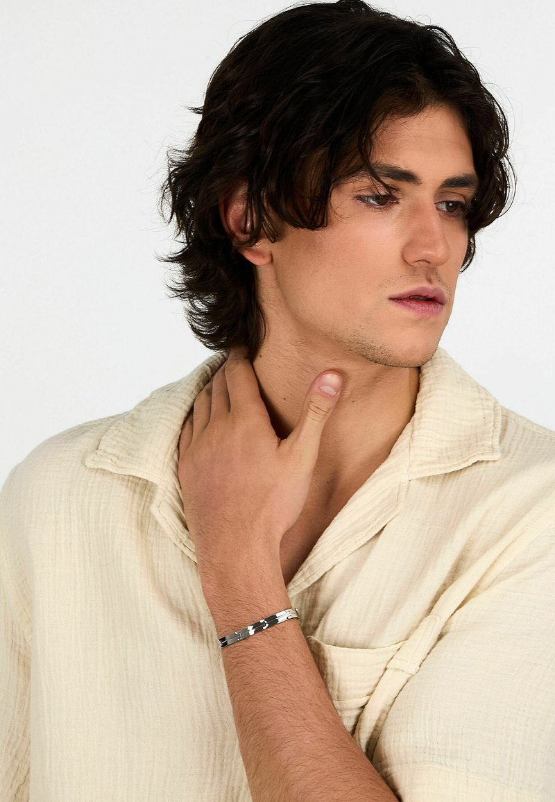 Bracciale in metallo argentato caratterizzato da un design elegante con accenti neri a contrasto, indossato su un polso contro una camicia chiara e strutturata.