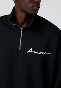 Sweat-shirt noir à demi-fermeture éclair avec une texture douce. Présente une broderie blanche "Amour" sur le côté gauche de la poitrine et une fermeture éclair en métal.