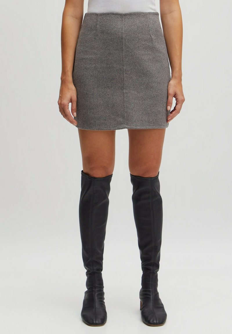 Femme portant une mini-jupe grise et des bottes noires montantes, debout devant un fond blanc uni.