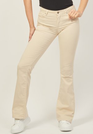 Pantaloni a zampa d'elefante beige realizzati in un mix di cotone, con vita aderente, tasche frontali e una texture liscia. Abbinati a sneakers bianche.