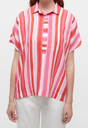 Vrouw die een kort mouws overhemd draagt met verticale roze, rode en witte strepen, gecombineerd met een witte broek, staand tegen een effen achtergrond.