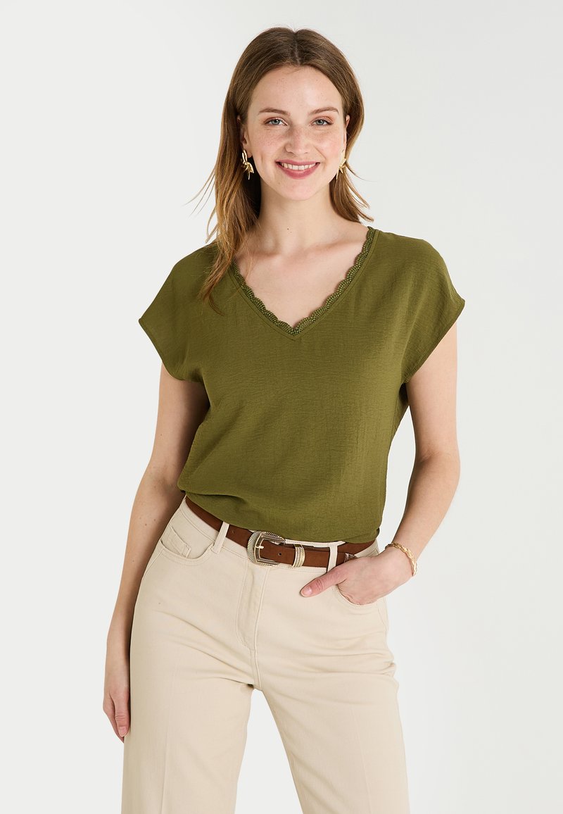 Haut vert olive à col en V avec des bords festonnés et des manches courtes, associé à un pantalon beige clair et une ceinture marron. Texture de tissu lisse.