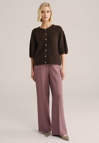 Barna rövid ujjú kardigán nagy gombokkal, magasderekú, bő szárú világos rózsaszín nadrággal és hegyes orrú bézs cipővel párosítva.