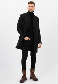 Cappotto in misto lana nero con chiusura a un bottone, indossato sopra un dolcevita nero, abbinato a jeans neri slim-fit e stivali stringati marroni.
