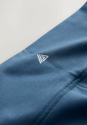 Niebieska tkanina sportowa o gładkiej fakturze z trójkątnym, białym logo i wzmocnionymi szwami. Widok z bliska, pod kątem.