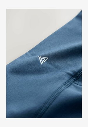 Le tissu sportif bleu présente une texture lisse avec un détail de logo blanc triangulaire et des coutures renforcées. Vue rapprochée, en angle.