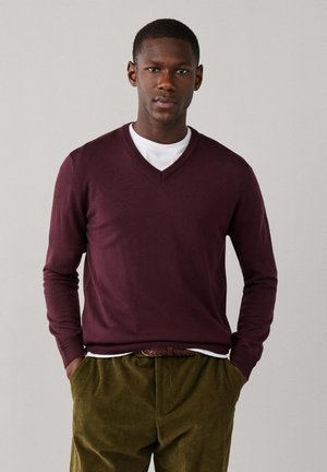 Hombre de piel oscura con un suéter de punto burdeos con cuello en V sobre una camisa blanca y pantalones de pana verde oliva, de pie con las manos en los bolsillos.
