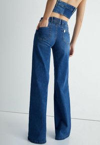Kvinna i högmidjade blå vida jeans och matchande denimcrop-top, stående med en hand i bakfickan mot vit bakgrund.