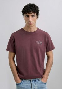 Camiseta de algodón marrón con mangas cortas, cuello redondo, que presenta un pequeño logotipo blanco en el pecho. Llevada con jeans de mezclilla azul desgastados.