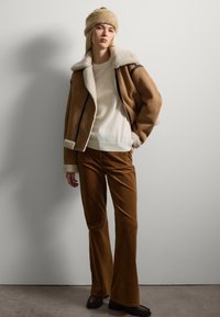 Bruine schapenvacht jas met crèmekleurige voering, gecombineerd met een witte trui en bruine corduroy broek. Zwarte schoenen maken de outfit af.