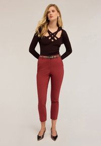 Svart ribbad tröja med utskärningar vid halsringningen, i kombination med åtsittande vinröda byxor och ett matchande bälte. Modellen bär svarta spetsiga skor.