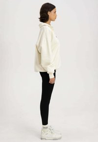 Overmaatse crèmekleurige hoodie, zwarte legging en witte sneakers met grijze accenten. Zachte textuur, casual design en zichtbare geribbelde manchetten.
