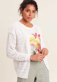 Weißer, gestrickter Cardigan mit Lochmuster, rundem Halsausschnitt und langen Ärmeln, getragen über einem bunten, floral bedruckten Hemd.