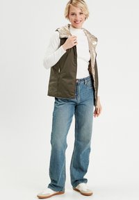 Gilet sans manches vert olive avec une capuche brillante de couleur claire, associé à un jean en denim bleu et des chaussures blanches. Le gilet est doté d'une fermeture éclair et de poches.