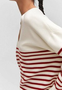 Nainen, jolla on letitetty poninhäntä, pukeutuu valkoiseen neulepaitaan, jossa on vaaleanpunaisia vaakaraitoja keskivartalolla ja hihoissa.