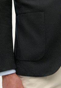 Blazer de pata de gallo oscuro con un ajuste estructurado, que presenta un bolsillo con solapa y un puño azul claro en contraste debajo. Textura suave.