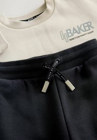 Felpa beige chiaro con logo "BAKER NINETEEN EIGHTY EIGHT", abbinata a pantaloni jogger neri con vita testurizzata e coulisse.