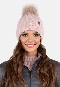 Bonnet tricoté rose clair avec un pompon duveteux sur le dessus, présentant une texture côtelée et une petite étiquette marron sur le côté.