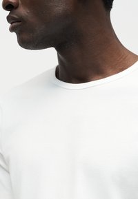 Gros plan d'une personne à la peau foncée portant un t-shirt blanc uni à col rond, tournée vers la gauche sur fond blanc.