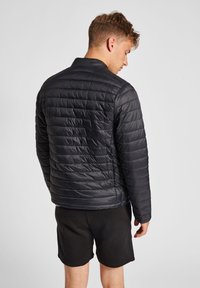 Svart, vadderad puffjacka med ståkrage och horisontella sömmar, bärs med svarta shorts. Slitstarkt, lätt material.