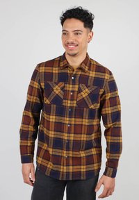 Chemise en flanelle à carreaux pour homme, dans des tons de bleu marine, moutarde et rouille. Dotée de manches longues, de deux poches poitrine et d'une fermeture à boutons. Texture de tissu douce.