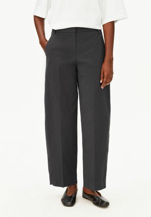 Personne portant un pantalon noir à jambes larges, des chaussures plates noires et un haut blanc à manches courtes, une main dans la poche.