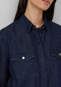 Dunkelblaue Denim-Hemd mit Button-down-Kragen, zwei Brusttaschen mit goldfarbenen Beschlägen und einer glatten Textur.