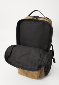 Brown canvas backpack with a black interior. Zippered opening reveals a spacious main compartment and additional pocket. Smooth texture.