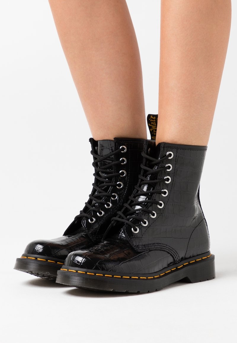 Bottes à lacets noires brillantes à motif crocodile portées sur des jambes nues, avec des semelles épaisses et des coutures jaunes.