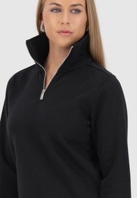 Pullover nero con zip a quarto di lunghezza e colletto alto, realizzato in tessuto morbido. Presenta una zip argentata e linee pulite per un look aderente.