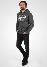 Grijze hoodie met witte logotekst, loose fit, voorzak, gecombineerd met zwarte gescheurde jeans en zwarte sportschoenen.