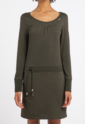 Kobieta ubrana w długą sukienkę z długim rękawem w oliwkowym kolorze, z dekoltem w kształcie litery U oraz regulowanym wiązaniem w talii.