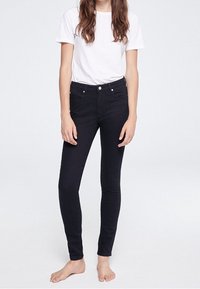 Jean skinny noir à taille haute, en denim. Comprend des poches avant discrètes et une texture lisse. Porté avec un t-shirt blanc uni.