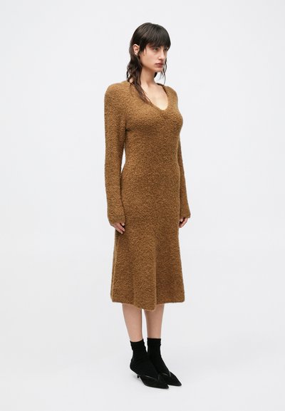 Bruine, textuurvolle jurk tot op de knie met lange mouwen en een V-hals. Draagt zwarte enkelsokken en puntige zwarte schoenen.