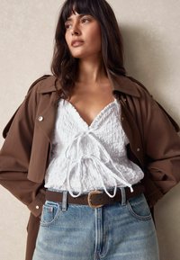 Haut blanc froissé avec détail à nouer, trench-coat marron, ceinture sombre et jean bleu clair avec une légère décoloration. Texture lisse et coupe décontractée.