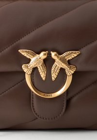 Καφέ δερμάτινη τσάντα χειρός με καπιτονέ σχεδίαση, χρυσό διπλό κούμπωμα με έμβλημα που απεικονίζει δύο πουλιά και ορατό όνομα μάρκας "PINKO" από κάτω.