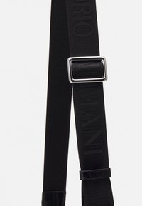 Svart justerbar vävd rem med silverfärgad rektangulär spänne och präglat varumärkesnamn på texturerad tyg.