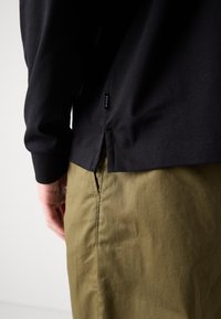 Fekete, hosszú ujjú póló oldalhasíték részlettel és "BOSS" felirattal, olíva zöld nadrággal viselve. Simaságával kiemelkedő anyag, hétköznapi stílus.