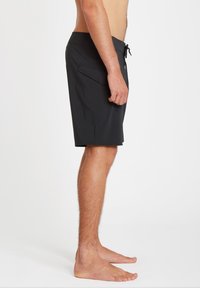 Homme debout pieds nus, portant un short de bain noir mi-cuisses avec poche latérale et taille à cordon, présenté de profil sur fond blanc.