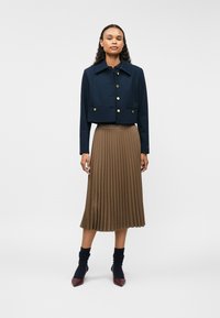 Jachetă scurtă bleumarin cu nasturi aurii, asociată cu o fustă maro plisată. Accesoriile includ șosete bleumarin cu model și pantofi maroon cu vârf ascuțit.