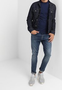 Mörk denimjacka, marinblå tröja med rund hals, figursydda blå jeans och grå sneakers. Anmärkningsvärda detaljer inkluderar metalldetaljer och uppvikta ärmar.