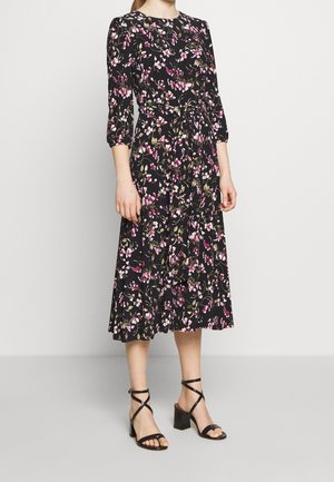 Robe noire à fleurs avec manches longues, ornée de fleurs roses et d'accentuations vertes, taille cintrée et jupe évasée. Associée à des sandales à talons noires.