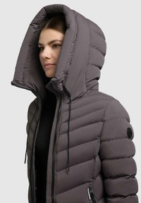Una donna che indossa un giubbotto imbottito grigio scuro con un ampio cappuccio, chiusura con zip e coulisse, mostrata di lato su uno sfondo neutro.