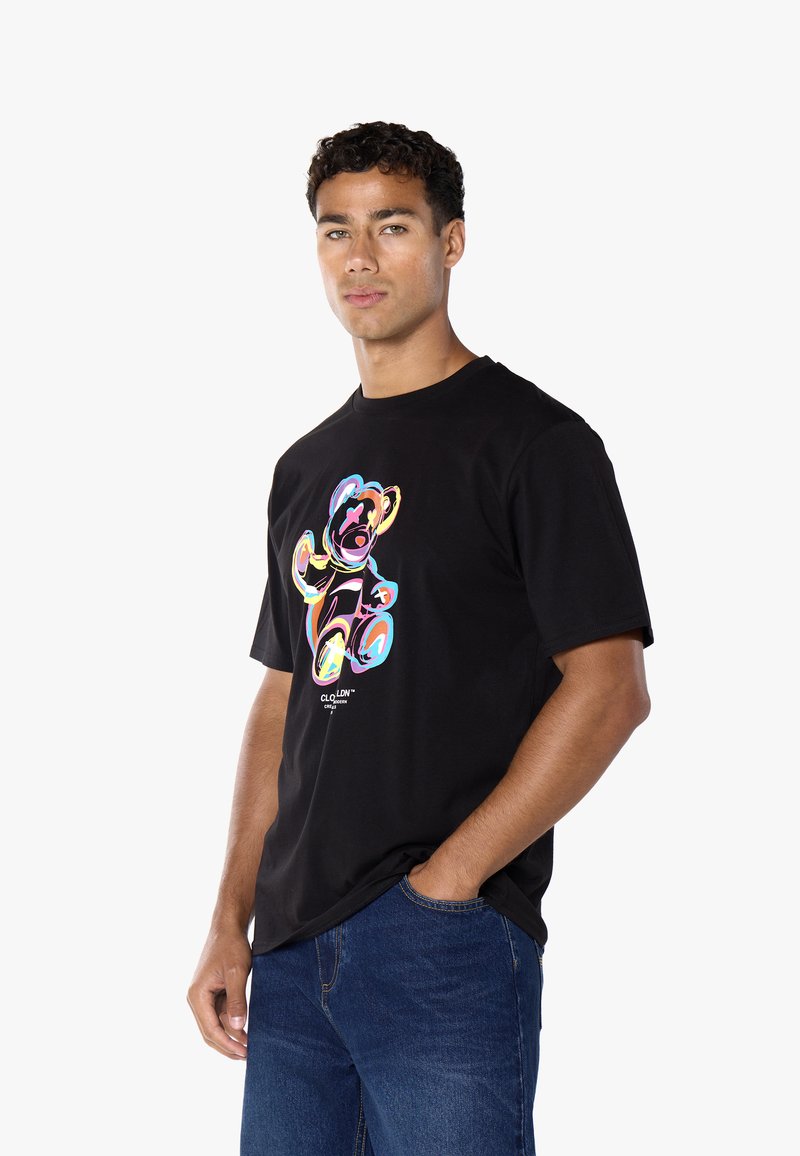Uomo con capelli ricci che indossa una maglietta nera con un design astratto colorato di orsetto e jeans blu, in piedi con una mano in tasca.