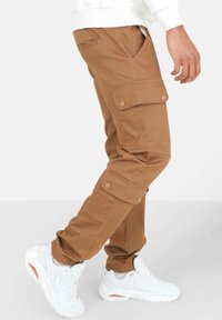 Bruine cargobroek met meerdere zakken, knoopsluitingen en een taperende pasvorm. Draag met witte sneakers met oranje accenten.