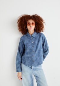 Blue textured button-up shirt with a collar, featuring a subtle pattern, paired with light blue jeans and round sunglasses.
