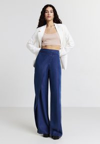 Donna che indossa un blazer bianco, un top corto beige e pantaloni navy a gamba larga con un pannello frontale sovrapposto, in piedi con le mani sui fianchi.